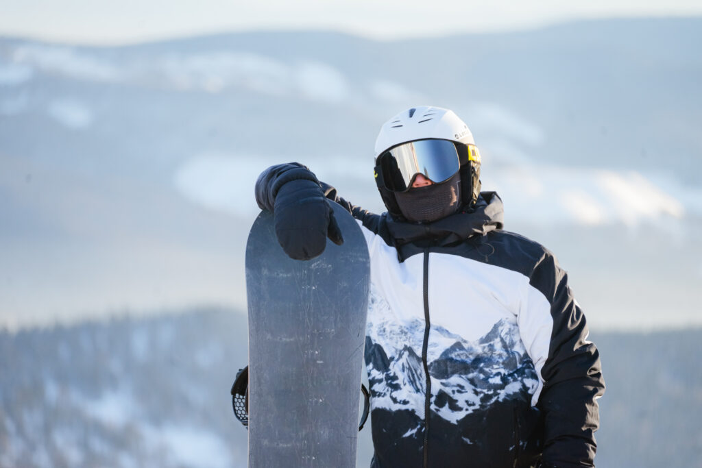 A close-up of a man wearing ski goggles and a helmet, standing on top of a mountain with a snowboard in his hands, looking at the camera.